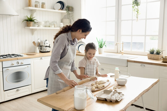 Family bonding through cooking. Happy loving young mom give culinary lesson to little schoolgirl daughter teach to knead roll dough help to use simple kitchen utensils to cook homemade bakery treats