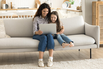 Digital education. Caring millennial mother babysitter sit on couch with little daughter junior...