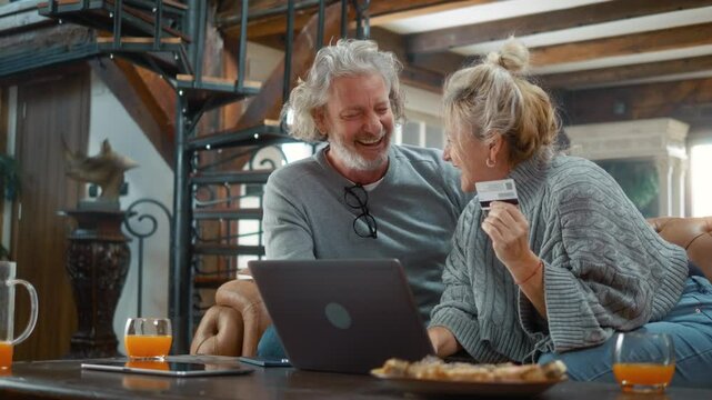 Happy, elderly couple in their 50s is at home using a laptop and credit card to make online payments, order, or book vacation reservations. They hug and celebrate a successful transaction.