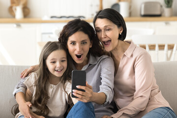Can you believe it. Friendly family older granny adult mom little girl hug on sofa scream in...