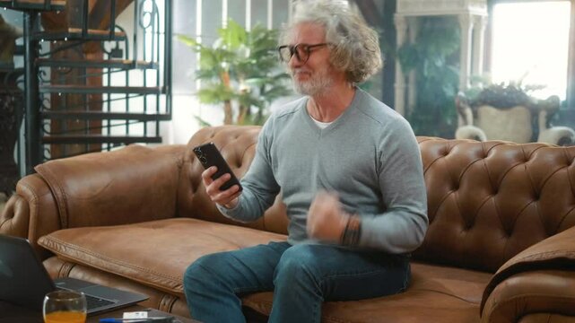 Surprised 60s elderly man gets good news message on mobile phone while sitting on couch at home, celebrate success mobile win online bet
