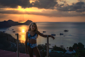 Fototapeta premium A young girl joyfully poses against a breathtaking sunset backdrop, overlooking the peaceful ocean and the distant silhouette of majestic mountains in the horizon, Komodo, Indonesia