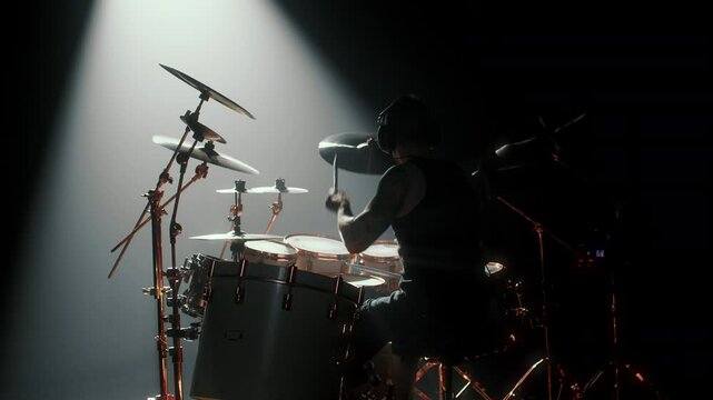 Silhouette of professional drummer on stage in darkness, gig in concert hall. Cinematic backlit view of male musician punching drum set, power of music and rhythm, back view, rock musical performance