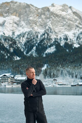 Fototapeta premium Contemplative Man in Black Outfit Admiring Snowy Alpine Landscape with Frozen Lake and Traditional Village. Serene Winter Mountain Scene Reflecting Tranquil Beauty of Nature in High Resolution