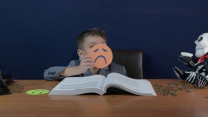 A boy with sad smiley over his face studdying finances. Coins and skeleton piggy bank on the desk....