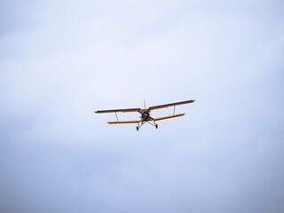A yellow vintage biplane captured mid-flight against a vast cloudy sky. The classic aircraft's front view showcases its wingspan and intricate details.