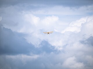 A small yellow biplane soars high in the sky, surrounded by dramatic white and gray clouds. The vast sky emphasizes the sense of adventure and freedom.