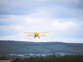 A vintage yellow biplane soars into the sky above a lush green landscape with distant buildings. The cloudy sky adds depth to the scenic aviation moment.
