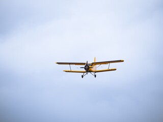 A yellow vintage biplane captured mid-flight against a vast cloudy sky. The classic aircraft's front view showcases its wingspan and intricate details.