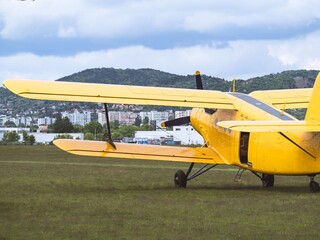 A classic yellow biplane parked on a grassy airfield with a cityscape and mountains in the background, showcasing vintage aviation and aircraft design.