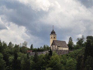 Fototapeta premium A historic church sits atop a lush green hill in Tarvisio, Italy, near the Alpe-Adria cycling route. The cloudy sky adds a dramatic touch to the serene and picturesque landscape.