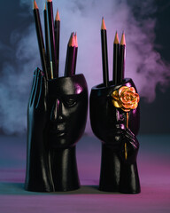 Two black plaster cups shaped as male and female faces, each holding several black pencils on a gray background with pink and blue highlights and smoke. 