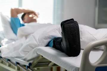 Patient resting in hospital bed, wearing walking boot, hand on forehead, displaying physical...