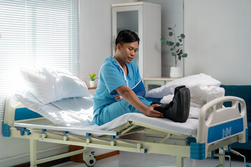 Asian patient recovering from injury, wearing arm sling and walking boot, seated on hospital bed...