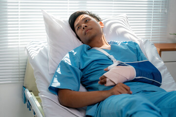 Young Asian male recovering from surgery, wearing arm sling, lying in hospital bed and gazing...