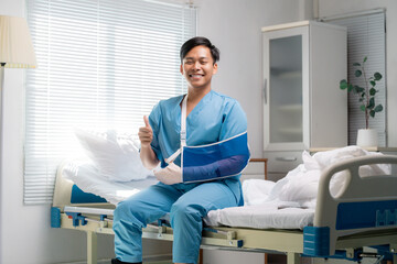 Healthcare professional in blue scrubs seated on hospital bed, wearing arm sling, giving thumbs-up...