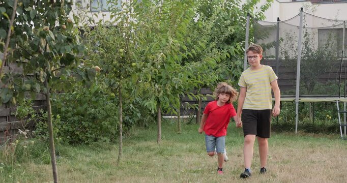 Little kid with fractured leg in gypsum plaster cast helped by his big brother to walk limping through the garden, front view