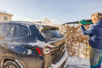 Man uses an electric leaf blower to remove snow from black SUV parked in driveway of suburban house...