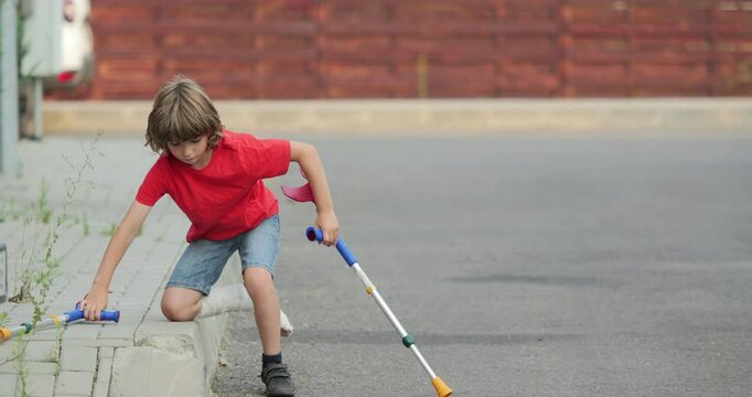 Child with a fractured leg in gypsum plaster cast gets up from the sidewalk border curb with the help of medical orthopedic crutches and walks on