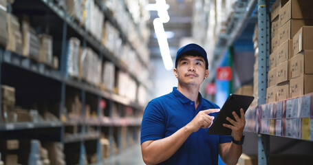 A man in a blue shirt is pointing at a tablet in a warehouse. He is wearing a blue hat and he is...
