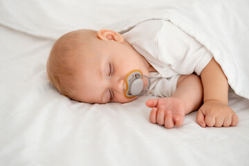 a newborn baby sleeps sweetly sucking a pacifier, a place for text, a baby sleeps on a white bed under a blanket with a pen under his cheek, a healthy baby's sleep