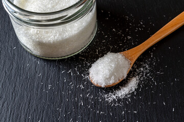 Monosodium glutamate in a wooden spoon and glass jar full of MSG white crystals over black...
