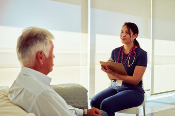 Fototapeta premium Friendly nurse taking notes while listening to senior man describing symptoms during medical consultation in bright hospital room