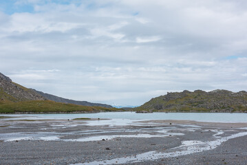 Rocky isle on blue mountain lake with stony sandy shore in rainy weather. Ripples on azure water surface of alpine lake in high mountains under cloudy gray sky. Snow covered mountain range far away.