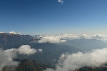 A stunning panoramic view of majestic mountain ranges covered in lush greenery, with white clouds drifting over the valleys. The distant snow-capped peaks contrast beautifully against the blue sky