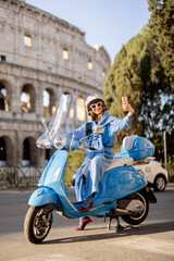 Stylish woman in a blue outfit and helmet captures a joyful selfie while sitting on a classic scooter in front of the Colosseum in Rome. A perfect blend of modern lifestyle and historic charm © rh2010