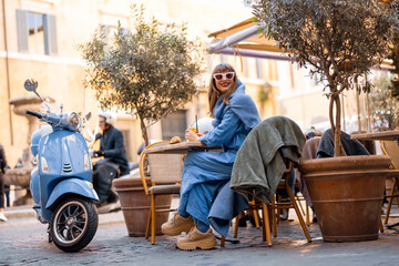 A fashionable Italian woman sits at a cozy outdoor cafe in Rome, sipping an aperitivo. Her scooter...