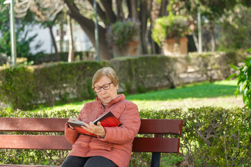 Obraz premium Senior woman sitting in a bench in the park, reading a book