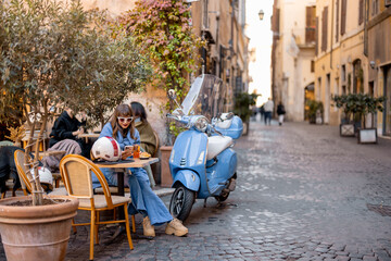 Plakat A fashionable woman enjoys aperitivo and a croissant at a charming Roman cafe. With her stylish outfit, sunglasses, and scooter, she embodies the essence of Italian elegance and relaxed city life