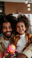 A joyful Black father and daughter are celebrating Easter together indoors, holding a brightly colored egg and smiling widely