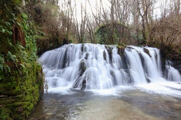 Paisaje de cascada de agua  efecto seda con molino de piedra al fondo y detalle en primer plano de muro de piedra con musgo 