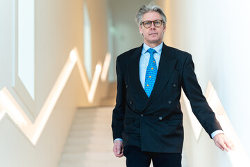 A serious businessman wearing glasses and a formal black suit stands before a simple light...