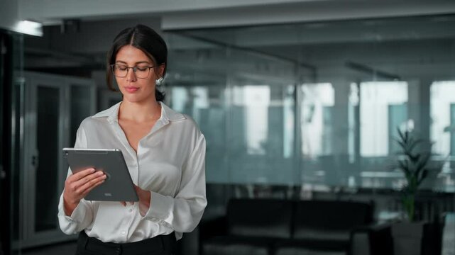 Hispanic mature professional business woman looking at pc and using app. European businesswoman CEO holding digital tablet working fintech tab application standing at workplace in office. Copy space
