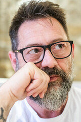 A 50-year-old man with glasses is seated alone at home, his expression full of sadness and...