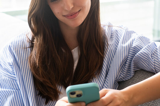 Modern millennial woman at home sitting on a sofa, dressed casually, scrolling on a smartphone screen, checking social media, chatting with friends, making new connections, reading web pages
