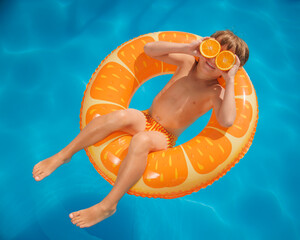 Happy child on an inflatable ring holds orange halves like sunglasses. Funny kid is relaxing in the swimming pool on summer vacation