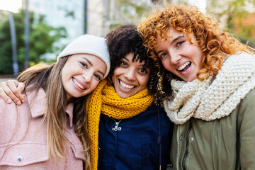 Three diverse women friends happily posing together on a cold winter day in the city, bundled up in...