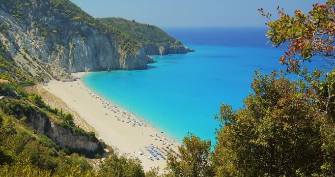The picturesque Mylos beach in Lefkada island, with turquoise waters of the Mediterranean sea washing the white pebbled shore, framed by high rock cliffs covered green lush vegetation and pine trees