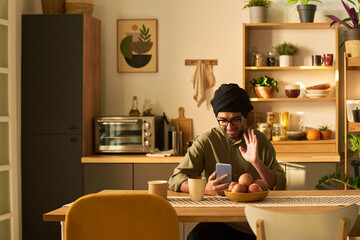 Man sitting in cozy kitchen, holding smartphone while touching beard and sitting at table with bowl of fruit. Warm ambient lighting highlighting kitchen decor and wooden elements