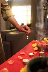 Hand holding brass bell near table decorated with various items. Table includes flowers, lit candles, and incense sticks in cozy living room setting