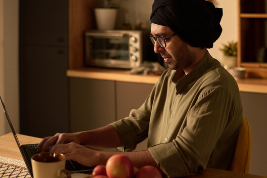 Man wearing a turban focusing on typing at a laptop. Background includes kitchen elements like microwave and fruit bowl on wooden counter