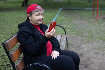 Portrait of happy elderly woman in red cap and sportwear sits on bench and smilingly talks on smartphone on background of green park. Active life of pensioners and adaptation in the modern world. Prev