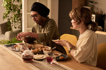 Two people sitting at dining table, savoring an array of Asian dishes under warm indoor lighting, engaging in relaxed dinner conversation