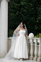 woman in a wedding dress. She stands against the backdrop of green bushes. The dress is white,...