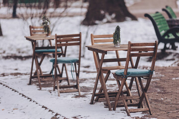Wooden tables and chairs sit on a snowy path, each featuring a small green plant centerpiece, creating a serene and inviting atmosphere in a winter park