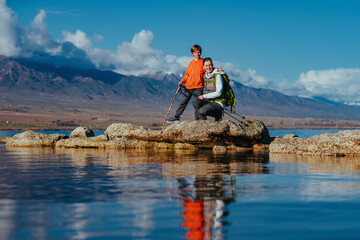 Mother and son tourists posing on stones on lake shore on high mountains background